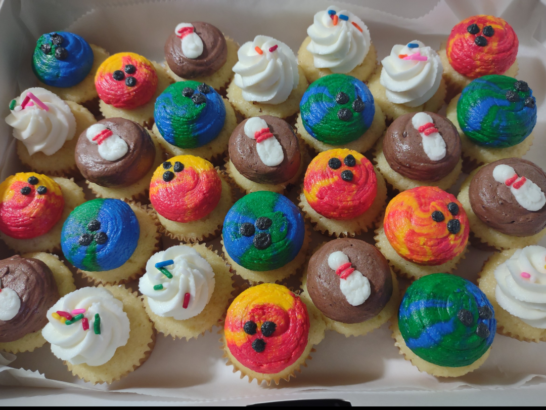 A variety of colorful cupcakes with different designs and frosting, displayed in a white box.