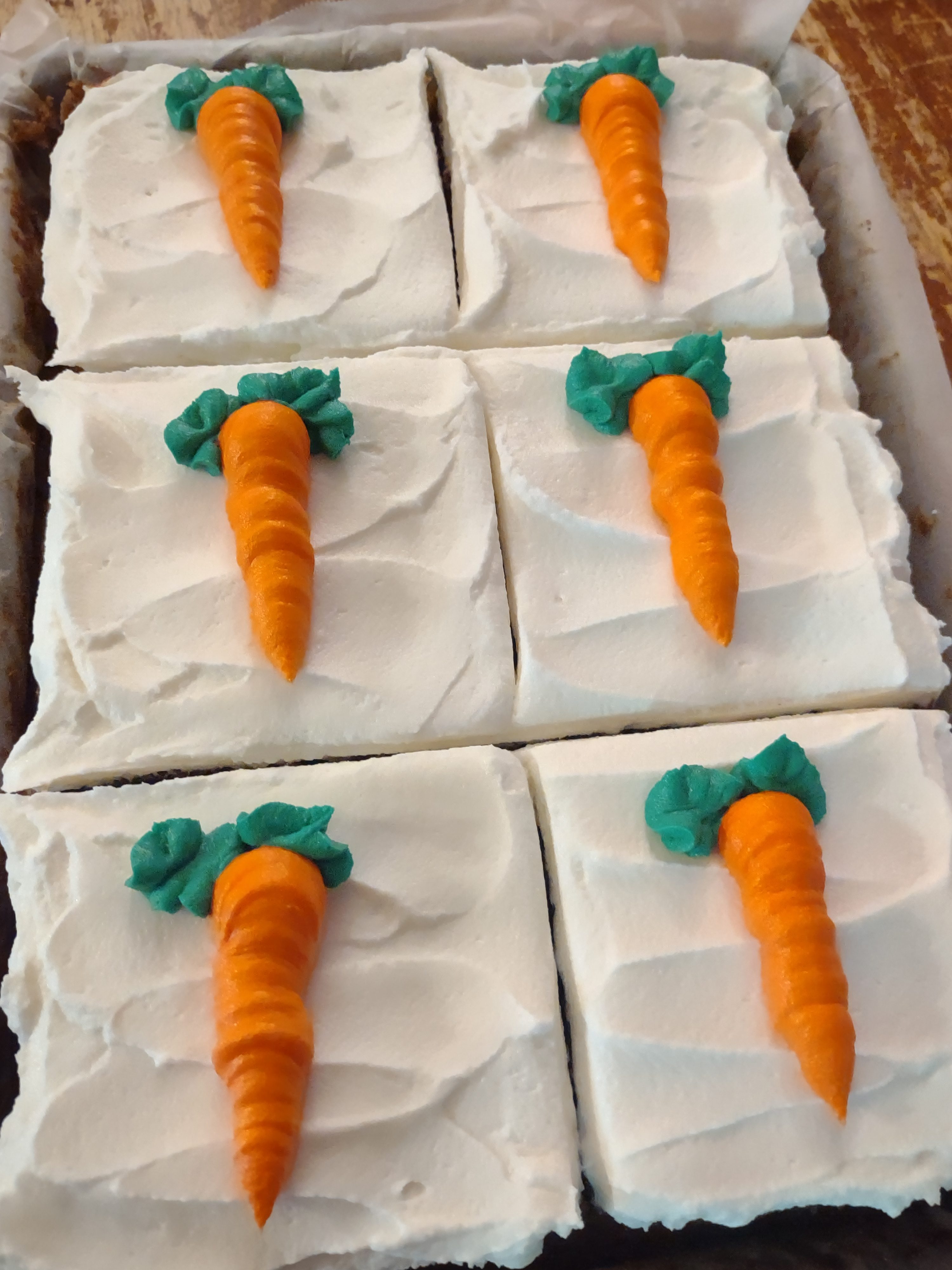 A tray of freshly baked sheet cakes topped with white frosting and adorned with colorful, decorative carrot toppers.